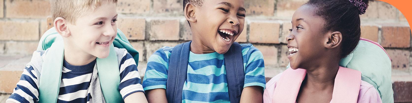 Na imagem temos 3 crianças sorrindo e olhando entre si, dois meninos na esquerda para a direita, e uma menina na direita. As três crianças estão utilizando mochilas